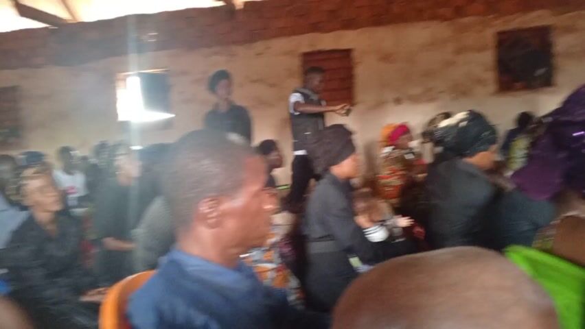 The video shows a group of people gathered in what appears to be a simple, possibly rural, building. The walls of the building are mostly unpainted brick, and the room is lit by sunlight coming through small windows. The roof appears to be made of wood and corrugated metal.  Several people are seated in chairs, while a group stands near the front of the room, singing and clapping. A man with a microphone seems to be leading the group. Most of the people are African. Some of the women are wearing traditional head coverings. The atmosphere seems festive or religious, with a sense of community and celebration.
