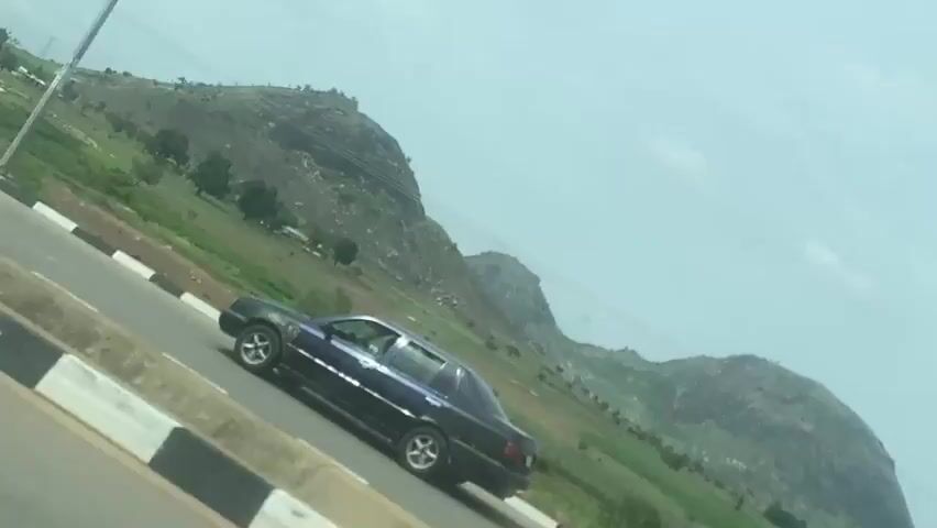 The video captures a daytime view from a moving vehicle, looking out onto a road and a natural landscape. A dark-colored sedan, possibly dark blue or black, is seen driving on the road, moving from left to right across the frame. The road features a concrete barrier on the left with distinctive black and white stripes. A tall, thin pole, possibly a utility pole or street light, stands next to the road. In the background, there are several large, rocky hills or mountains covered in green vegetation, set against an overcast or hazy sky. The environment appears to be an open, possibly rural or semi-urban area with lush greenery. No specific activity beyond the car driving is depicted, and no visible text can be discerned. The scene is consistent with the provided context of Gosa, Nigeria.