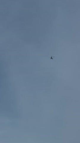 The video shows a helicopter flying in a blue sky with some thin clouds. The helicopter is a light color, possibly white or silver, and appears to be a smaller model. It is seen from below, moving across the frame at a distance.