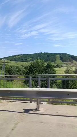 The video shows a scenic landscape from a moving vehicle. A guardrail runs horizontally across the foreground, blurred by motion. Behind the guardrail, a vibrant green field stretches towards a gently rolling hill covered in dense green trees. A ski slope is visible on the side of the hill, with some areas cleared of trees. The sky above is a bright blue with scattered white clouds. A single utility pole stands in the middle ground of the field. The overall impression is one of a clear, sunny day in a rural, hilly environment.