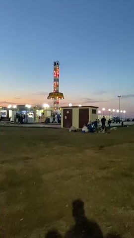 The video shows an outdoor amusement park scene at dusk. A tall, brightly lit drop tower ride is the central focus, with its lights cycling through various colors. In the foreground, a grassy field is visible, with the shadows of two people cast on the ground. To the left of the drop tower, there are several food stalls or game booths with lights illuminating them, and a few people can be seen standing around these areas. To the right of the drop tower, there are two small, attached buildings, one with a tan facade and a red door, and another with a red facade and a tan door. Several people are sitting or standing in front of these buildings, some appearing to be resting or socializing. In the background, the sky is a gradient of blue and orange, indicating sunset, and a few more lights are visible in the distance, suggesting more structures or lights beyond the immediate scene. The overall impression is of a lively but relaxed evening at a temporary amusement park.