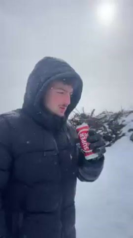 Here is a brief description of the video:
The video shows a person in a snowy environment. The individual, wearing a black puffer jacket with a hood and gloves, drinks from a red and white can labeled "SUPER." The sun shines weakly through a cloudy sky. After taking a sip, the person turns away from the camera and walks a short distance up a snow-covered slope, which leads to a view of a valley shrouded in fog. A few sparse bushes are visible in the foreground. The scene is overcast with light snow falling.