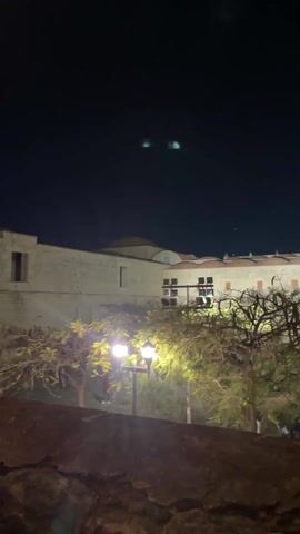 The video shows a building at night, partially obscured by trees. The building is lit up and has two bell towers. There are streetlights in front of the building. Some people are walking around. The video pans up to reveal more of the building.