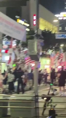 The video shows a group of people walking on a street at night. Several people are holding flags, including the flags of South Korea and the United States. Some of the flags are large and on poles, while others are smaller and held by individuals. There are businesses with illuminated signs in the background, suggesting an urban setting. The people appear to be moving in the same direction, possibly in a protest or demonstration. The lighting is dim, and there is some motion blur, indicating that the video was likely recorded from a moving perspective.