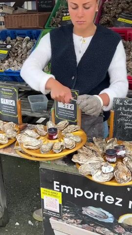 A woman with dark hair tied back, wearing a white sweater and a dark blue vest, is working at an outdoor oyster stand. She is focused on shucking oysters, holding an oyster in her left gloved hand and using an oyster knife in her right hand. On the wooden table in front of her are several yellow, round trays filled with shucked oysters. Small jars with red contents are placed on these trays. To her right, there are more oysters on display. Behind her, various crates overflowing with oysters are visible. Signs with French text and prices are displayed on the stand.