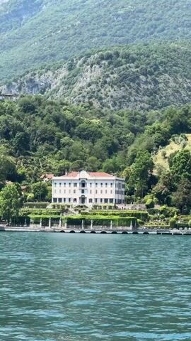 The video shows a lake with a building in the background. The building is white with a red roof and is surrounded by trees and greenery. A mountain is also visible in the background. The water is a greenish-blue color. The camera is shaking slightly, suggesting it is handheld. The day appears to be sunny.