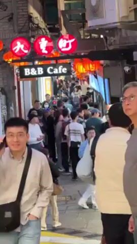 The video shows a crowded alleyway in an urban area. The alley is lined with buildings and decorated with red lanterns. Above the entrance to the alley is a sign that reads "B&B Cafe" with Asian characters above it. A large crowd of people are walking through the alley. Some people are looking at their phones, while others are talking to each other. There are several people looking at the camera.  The overall impression is of a busy tourist destination.