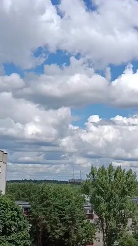 A time-lapse video shows a bright blue sky with puffy white clouds drifting by. Below the sky, a line of green trees forms a dense canopy. Behind the trees, several multi-story apartment buildings are visible, with some showing reddish-brown accents on their facades. In the far distance, two tall, slender industrial smokestacks with grey tops rise above the treeline. The video captures the movement of the clouds and the subtle shift in light, suggesting the passage of time on a clear day.