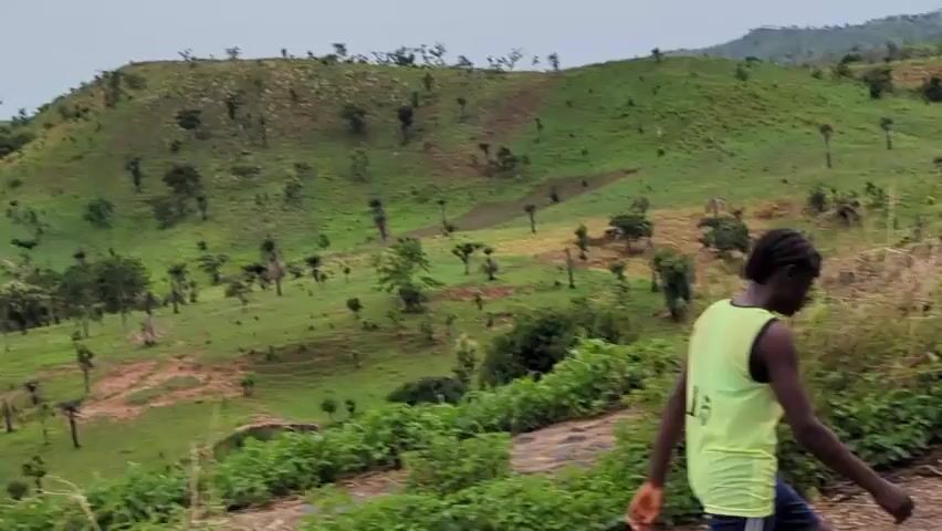 The video shows a young Black girl walking and then running uphill on a road. She is wearing a light green sleeveless shirt and black shorts with white stripes. The hillside behind her is mostly green but with patches of bare brown earth, and scattered small trees. In the distance, there is a hint of water. The sky is overcast. An unidentified person speaks in the background.
