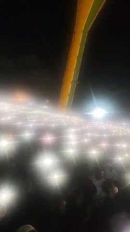 The video shows a nighttime outdoor event with a large crowd of people. Many individuals are holding up their phones, with the flashlights illuminating the scene, creating a sea of white lights. In the background, a tall structure, possibly a flagpole with a flag, is visible against the dark sky. There are also bright lights, including a prominent circular light source, contributing to the atmosphere. The overall impression is that of a gathering, possibly a concert or rally, with a festive or commemorative mood.