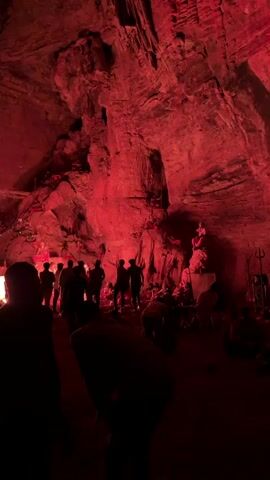 A group of people stand in a cave illuminated by red lights, observing a figure seated on a rock formation. The cave walls and ceiling are visible, textured and uneven, and the lighting casts deep shadows. The people in the foreground are largely silhouetted, with a few recognizable individuals among them. The central figure appears to be dressed in red and white, with a headdress. The overall impression is of a staged performance or exhibit within a natural cave setting.