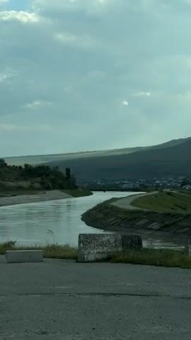 The video shows a serene landscape with a river winding through it. The river has a calm, reflective surface, mirroring the cloudy sky above. On the right bank of the river, there's a paved path or road, and further in the distance, a small town or village is visible nestled at the foot of rolling green hills. The hills are covered in lush vegetation and appear to be quite extensive. The overall atmosphere is tranquil and natural, with a hint of human settlement integrated into the landscape. The lighting suggests it might be late afternoon or early evening, with the sun partially obscured by clouds.