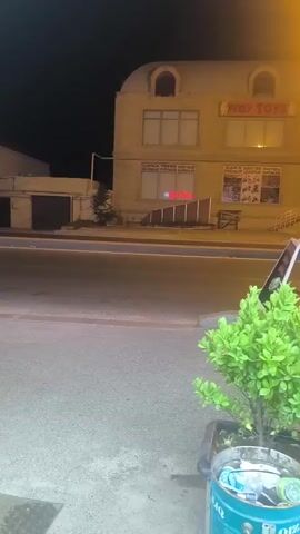 The video begins by showing a building at night. A sign on the building reads "Play Toys." A second sign reads "My Folder." To the left of the building, there is a dark garage door. In front of the building, there is a street and a sidewalk. A small green bush is in a planter on the sidewalk. To the right of the bush, a motorcycle is parked. The motorcycle has a red delivery bag on the back. The bag has a picture of a burger on it and reads "YANGO." The video pans to the right, showing more of the street and sidewalk. There is a streetlamp illuminating the area. The video ends showing the motorcycle and bushes from a slightly different angle.