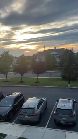 The video shows a suburban street scene at dusk. A young person on an electric scooter rides on a sidewalk past a street lamp and a real estate sign. Several parked cars are visible in the foreground. Houses and trees line the street in the background, and the sky is filled with dramatic clouds illuminated by the setting sun.