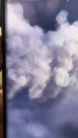 The video appears to be shaky footage, possibly taken from a handheld device. It begins with a shot of clouds, then transitions to a scene with dark smoke rising into the sky. A street sign is visible, and below it, a bridge or elevated roadway with people on it. The scene suggests a fire or explosion. The footage then pans out to reveal an industrial area with many cars parked. Finally, a distant view shows a hazy cityscape with some tall structures, and a small dark object, possibly an aircraft, is visible in the sky. There are watermarks or text overlays on the video.