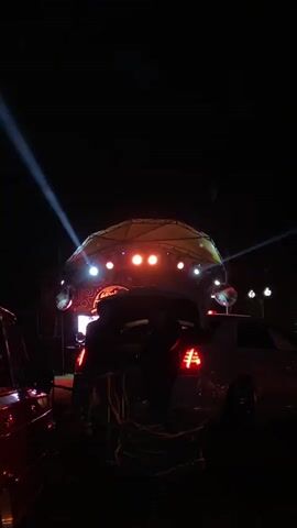 The video shows a nighttime outdoor event. In the foreground, there are parts of two cars visible, one on the left appears red and the other on the right is partially obscured but seems lighter in color. Between the cars, the rear of a dark-colored vehicle is prominent, with its hatchback open. Two people are silhouetted in the opening of the hatchback, with one appearing to be interacting with something inside. The rear lights of this vehicle are illuminated. In the background, a raised structure with a hexagonal canopy is visible, illuminated by multiple orange lights. Two bright white spotlights emanate from behind this structure, casting beams into the dark sky. Two circular objects, possibly speakers or lights, are mounted on either side of the structure. The overall atmosphere is dark, with the lighting focused on the central stage area and the cars in the foreground.