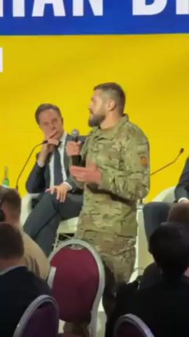 A man in camouflage stands at a podium speaking into a microphone. He has a beard and short hair. To his left sits another man in a suit. In the background is a yellow screen with white lettering. The word SCHOOF is partially visible on the podium. There are other people seated in the audience.