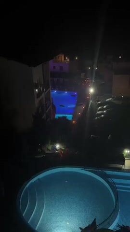 This video captures a nighttime view of a resort or hotel complex in Sozopol, Bulgaria. The scene is illuminated by artificial lights, giving it a distinctly nocturnal ambiance. The primary focus is on a large, brightly lit swimming pool with a deep blue hue, suggesting underwater lighting. Within the pool, a dark, elongated object is visible, appearing to be a decorative element or possibly a large inflatable toy, resembling a shark or some other marine creature. Another smaller, oval-shaped inflatable is also floating in the pool.

In the foreground, a circular section of another, smaller swimming pool is visible, also illuminated with blue light. The surrounding area appears to be landscaped with dark foliage, some of which are palm-like fronds. Beyond the main pool, there are buildings with illuminated windows, hinting at accommodation or other facilities. A row of parked cars is visible along a pathway, also lit by lampposts.

The overall impression is one of a tranquil, yet inviting, nighttime resort setting. The lighting creates a sense of artificial twilight, and the absence of visible people suggests a quiet time, perhaps late evening or early morning. The location cues point to a coastal resort area in Sozopol, Bulgaria, given the palm-like plants and the resort-style architecture. There is no visible text in the video. The scene evokes a feeling of relaxation and leisure, typical of a vacation destination.