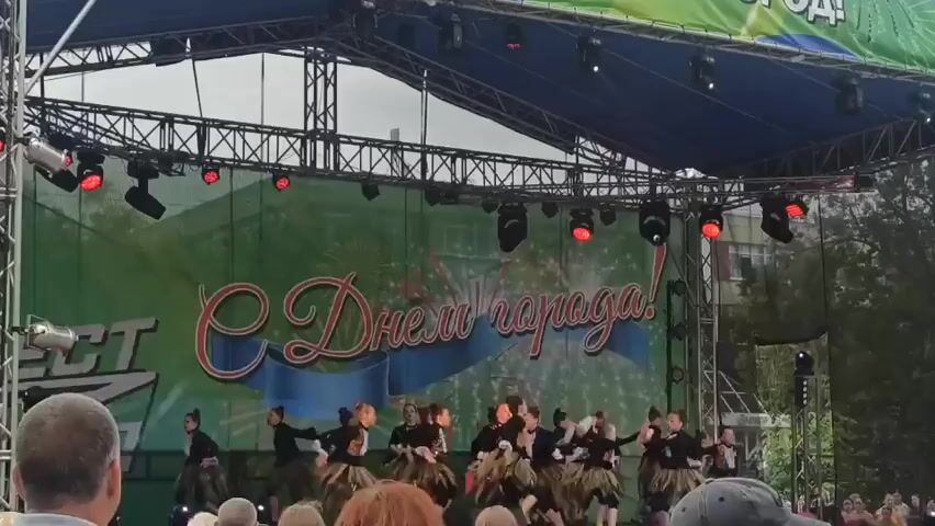 A group of young dancers in dark outfits with brown, fringed skirts are performing on an outdoor stage. The stage has a blue canopy supported by metal truss structures, with red spotlights visible. Behind the dancers, a large banner displays "С Днем города!" (Happy City Day!) in large, ornate red lettering, superimposed on a green background with fireworks imagery. To the left of the banner, a partial logo is visible with the word "PECT" in large white letters. In the foreground, the backs of several audience members' heads are visible, indicating they are watching the performance. The sky appears overcast.
