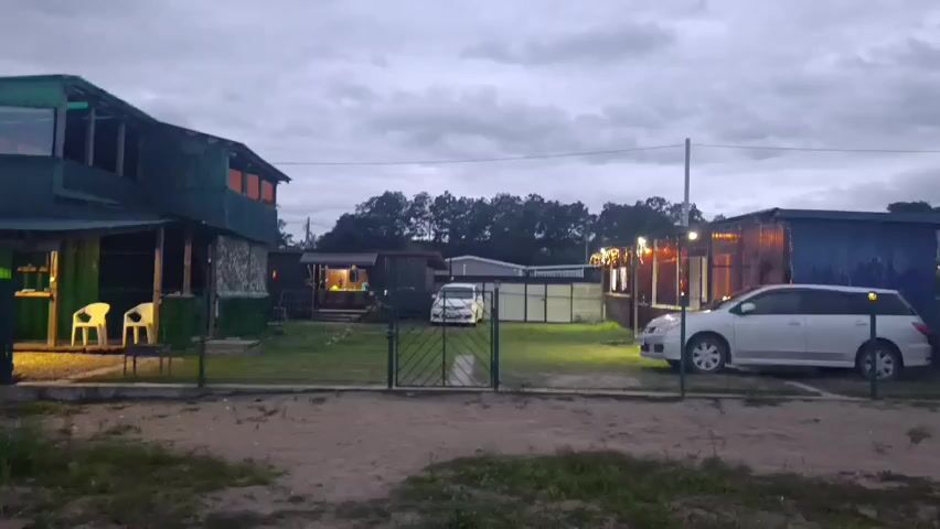 The video shows an exterior shot of a small complex of buildings at dusk. The sky is overcast. There are two white chairs and a table visible outside a green building on the left. A white car is parked to the right of a gate in the center of the frame. Another white car is parked in front of a blue building on the right. Lights are on inside some of the buildings, and string lights are visible above the fence on the right. There is a grassy area in front of the buildings, and a dirt path in the foreground. Trees are visible in the background. The overall impression is that of a rural or semi-rural setting.