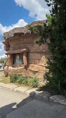 The video shows the exterior of a gas station with a prehistoric theme. The structure resembles a cave or rocky outcrop, with the ceiling made of what appears to be artificial rock formations and columns that look like oversized bones. A few people are visible, including a man and woman with a child standing near a vending machine and another person by a car at a gas pump. Trees and greenery surround the area, and a bright sky with some clouds is overhead. The ground is paved concrete.