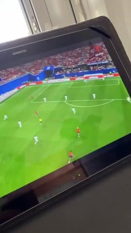 The video shows a tablet device displaying a soccer match on a screen. The game is viewed from a high angle, showing a green soccer field with players in white and red uniforms. The spectators in the background are wearing red and are seated in a stadium. The tablet is placed on a dark surface, possibly a desk or table. The top portion of the screen shows the brand name "HUAWEI". The game appears to be live, indicated by the FIFA World Cup branding on the screen. The action on the field involves players moving the ball and maneuvering for position within the penalty box. The lighting suggests it is daytime, with natural light coming from the left, casting a slight glare on the tablet screen. The overall scene is casual, implying someone is watching the game on their tablet.