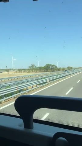 The video is filmed from inside a vehicle, likely a bus, looking out the window at a highway. In the distance, several wind turbines are visible against a clear blue sky. The highway stretches into the distance with cars driving on it. The scenery outside includes fields, some trees, and a guardrail separating the lanes. The interior of the vehicle shows a window, part of a seat, and a curtain. The video captures the motion of the vehicle as it travels along the highway.