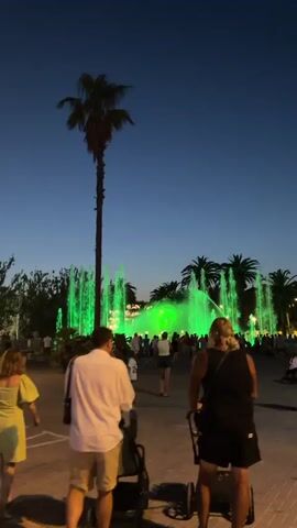 A vibrant fountain show takes place at dusk in Salou, Spain. The water features are illuminated with bright green lights, creating a dazzling spectacle against the darkening sky. Silhouetted palm trees frame the fountain, adding a tropical ambiance to the scene.

A diverse group of people are gathered to watch the display. In the foreground, three individuals are walking away from the camera. One person is pushing a stroller, another is carrying a bag over their shoulder, and the third is on a personal transport device. Further back, a crowd of onlookers are positioned in front of the fountain, some holding up their phones, presumably to capture photos or videos of the illuminated water.

The time of day is clearly evening, with the sky transitioning from day to night. The warm glow of the fountain suggests a pleasant, possibly warm, weather condition, conducive for outdoor evening entertainment. The presence of palm trees and the general atmosphere strongly indicate a Mediterranean coastal location, consistent with the provided location context of Salou, Spain. The mood appears to be one of relaxed enjoyment and shared community experience, as people gather to witness the impressive light and water show. No discernible text is visible in the media.