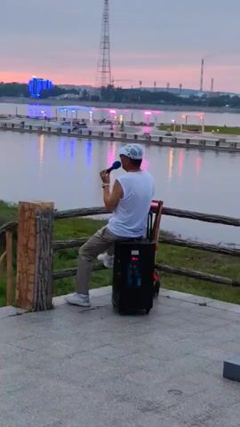 A man wearing a white sleeveless shirt, beige pants, and a white baseball cap sits on a black portable speaker with wheels. He is holding a blue microphone to his mouth and appears to be singing or speaking. He is facing away from the camera, looking towards a body of water. Behind him, across the water, is a city skyline with buildings, a tall tower, and industrial structures. The sky is twilight, with pink and purple hues. A wooden railing is in the foreground.
