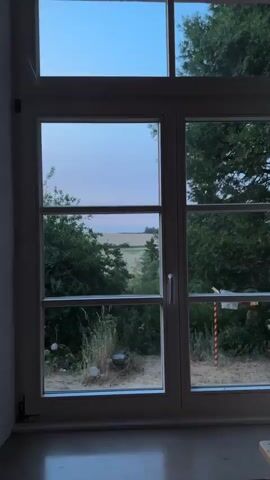The video opens with a view through a window showing a landscape with trees and a field under a pale sky. The camera then pans to the right, revealing the interior of a room with furniture. Specifically, the second and third frames show a sofa, a coffee table with two lit candles, and artwork on the wall. The lighting in the interior is dim, creating a cozy atmosphere. The final frame provides a closer view of the living area, highlighting the sofa, the coffee table, and the artwork on the yellow wall.