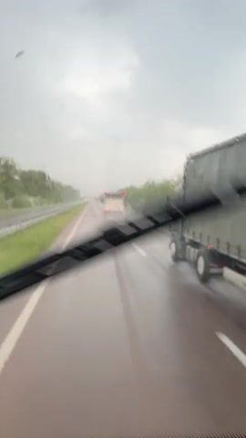 The video shows a view from inside a vehicle on a highway during a rainstorm. The road is wet and reflects the overcast sky. There are trees and greenery on either side of the highway, separated by a guardrail on the left. Several vehicles are visible ahead on the highway, including a white bus with red accents. The vehicle's windshield wipers are intermittently sweeping across the view, creating blurred streaks. The overall impression is of travel under adverse weather conditions.