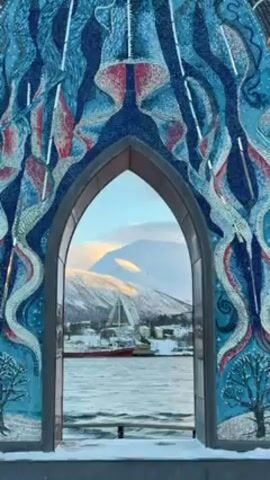 The video shows a stone archway with a mosaic on its surface, framing a picturesque landscape of a harbor, snow-covered mountains, and a city in the background. The mosaic features an abstract design with blue, red, and white tiles resembling waves or stylized northern lights, with small trees at the base. The archway is topped with a pattern of red and brown leaf-like shapes. There is snow on the ground around the arch. Through the arch, the water is visible along with ships, buildings, and a prominent mountain range bathed in the soft light of the day. Voices can be heard in the background.