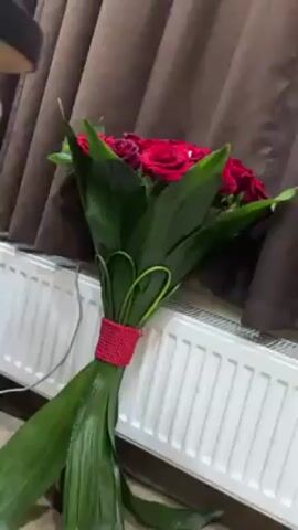 A bouquet of red roses and green foliage with a red beaded wrap around the stems leans against a white radiator in front of a brown and white curtain. The bouquet is slightly off-center, and the camera slowly zooms in and then out again. Cables and part of a dark chair are visible in the periphery.
