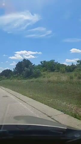 The video shows a view from inside a vehicle, looking out the windshield. The vehicle is traveling on a paved road with a white line dividing the lanes. To the right of the road is a grassy embankment with trees and foliage. The sky is blue with scattered white clouds. The scene appears to be a daytime outdoor setting, likely a rural or suburban area, with the vehicle in motion.
