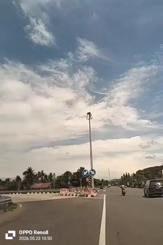 The video shows a view from a vehicle as it drives on a paved road. There are white lane markings on the road and a guardrail separating the lanes. A white van is traveling in the opposite direction. The sky is partially cloudy with a blue tint. Palm trees and other greenery line the side of the road. In the first frame, the vehicle approaches a roundabout with directional signs and a tall light pole. A motorcycle and a car are also visible on the road. The timestamp at the bottom reads "OPPO Reno8, 2026.05.23 10:50."