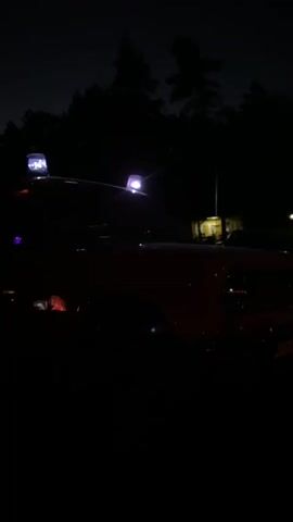 A red vehicle is parked at night in an outdoor location with trees in the background. The vehicle has two flashing lights on its roof, one blue and one white, and its headlights are on. The scene is dimly lit, with only the vehicle's lights and a distant light source illuminating the area.