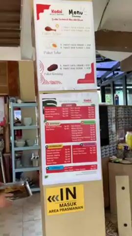 Here is a description of the video:

A bright and upright menu is displayed in what seems to be a casual dining establishment. The menu is divided into three sections, each with its own header. The top section lists "Paket Ayam" (Chicken Packages), "Paket Telur" (Egg Packages), and "Paket Rendang" (Rendang Packages). Each package contains a different combination of dishes, with the prices clearly listed. Below this, there is a section titled "Ala Carte Prasmanan" (A La Carte Buffet), detailing various individual food items such as "Ayam Goreng" (Fried Chicken), "Ayam Bakar" (Grilled Chicken), and sides like "Tahu Telur" (Tofu Egg). The prices for each item are also provided. The third section features "Gorengan" (Fried Snacks) like "Tempe Mendoan" and "Bakwan Jagung", as well as "Ice Cream" options like "Ice Cream Cone" and "Donat Ice Cream". Again, prices are listed for each item. A yellow sign with black lettering indicates "MASUK AREA PRASMANAN" (Enter Buffet Area).

A woman wearing a red shirt is seen standing next to the menu, appearing to be an employee of the establishment. In the background, there are additional people, what appears to be a buffet setup, and an "OUT" sign. The setting has a bright and simple decor, suggesting a casual and informal atmosphere.