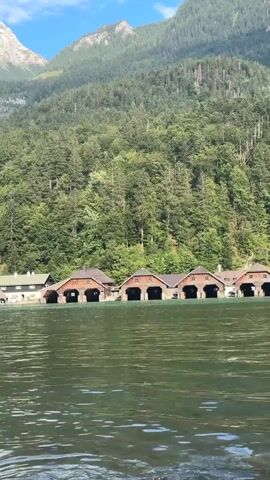 The video shows a body of water in the foreground with ripples. In the middle ground, a row of boathouses with arched entrances is visible along the shore. Behind the boathouses, a dense forest covers a steep mountainside, and in the background, a mountain peak rises against a blue sky. The scene appears to be a serene, natural landscape with man-made structures integrated into it.
