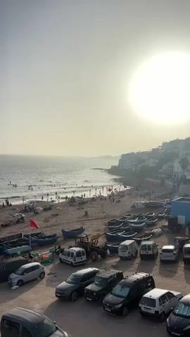The media file depicts a bustling beach scene in Taghazout, Morocco, during what appears to be a sunny afternoon. The vast expanse of the ocean dominates the left side of the frame, with waves gently rolling onto a sandy shore populated by numerous people. These individuals are engaged in various beach activities, including swimming, sunbathing, and strolling along the water's edge.

The foreground is a sandy area filled with parked vehicles, including cars and a tractor. Several blue fishing boats are lined up on the sand, some covered and others exposed, suggesting a working fishing community. Red flags are planted in the sand near some of the boats. Beyond the beach, a town or village with white buildings is visible on a hillside, overlooking the coast. The sky is bright and hazy, with the sun prominently featured, indicating it's likely late afternoon. The overall atmosphere appears lively and relaxed, capturing the essence of a popular coastal destination.

A sign with Arabic text is partially visible near a cluster of boats. The visible text reads "نشاطات إحساس" which translates to "Sensation Activities".