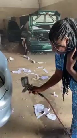 In a garage-like setting, a woman with long black dreadlocks stands on the right side of the frame, dressed in a blue floral-patterned shirt and beige pants, looking down at a car she is holding a paint sprayer towards. The floor is grey and covered with crumpled white paper. Behind her is a green car with its hood and trunk open, showing damage. The woman has her right hand on her neck and appears to be in discomfort, possibly coughing. The room is not well-lit, and the scene suggests an auto repair shop.