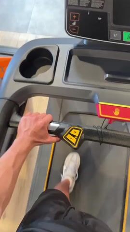 This is a first-person perspective video of someone on a treadmill at a gym. The video begins with a close-up of the person's hand gripping the treadmill's handlebar, and their feet are visible on the moving belt. The treadmill's console displays a speed of 1.5 mph. The camera then pans up to show more of the console, including various buttons and a digital display. The video then shifts to a wider view of the gym environment, showing other people in the background, exercise equipment, and architectural details like exposed beams and lighting fixtures. The overall impression is that of a typical gym setting.