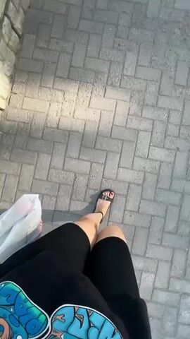 A first-person perspective video shows a person walking on a paved path. The person is wearing black shorts and black sandals. They are carrying a white plastic bag in their left hand. The path is made of grey rectangular pavers arranged in a herringbone pattern. To the left of the path is a low wall constructed of rough-hewn stones. Sunlight casts shadows on the path.