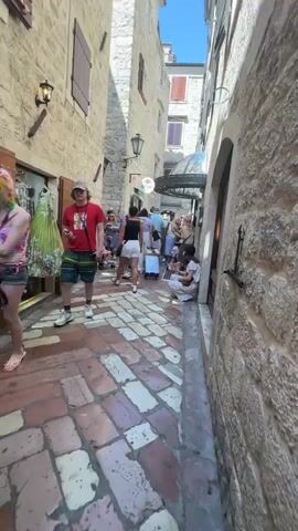 The video depicts a narrow cobblestone alleyway with stone buildings on either side, suggesting an old European town. The alley is populated with a crowd of tourists, many of whom are dressed in casual summer clothing and are pulling wheeled luggage. The sun is shining, casting distinct shadows, and the sky is blue and clear. In the foreground, a young person in a red t-shirt and patterned shorts is walking towards the camera. Further into the alley, several individuals are walking in various directions, some interacting with each other. One person is sitting on the ground against a building wall, holding what appears to be a piece of food. The buildings feature traditional stone construction, with some windows and awnings visible. Signs for a business named "SENSO" are posted on the walls. The overall impression is of a busy tourist area during daylight hours.