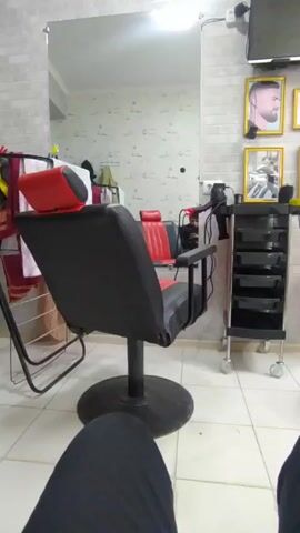 The video shows an interior shot of what appears to be a barber shop or hair salon. A black and red barber chair is in the center of the frame, facing a large mirror that reflects the room and other items. To the right of the chair is a black rolling cart with several drawers. The wall is decorated with a brick-like pattern and has framed pictures, including one of a man with a beard. A television is mounted high on the wall. A garment rack with various items of clothing and towels is visible on the left side of the frame. The floor is tiled. The lower portion of a person’s legs, clad in black clothing, is visible in the foreground, suggesting the video is taken from a seated position.