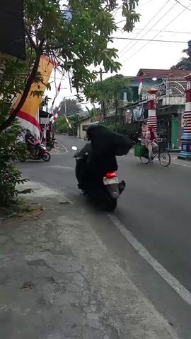 A scooter carrying two passengers, one wearing a black head covering and the other a dark jacket, turns left onto a street. A second scooter with a rider wearing a red shirt approaches from the opposite direction. Further down the street, a third scooter carrying a person with a green basket on the back is visible. A woman on a bicycle with a green basket attached is also seen riding on the right side of the street. The scene appears to be a street in a residential area, with buildings lining both sides of the road. Indonesian flags are displayed along the left side of the street. The sky is overcast.