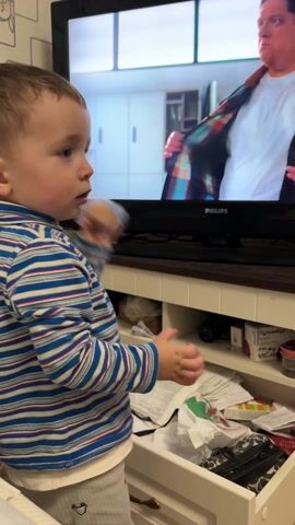 A toddler stands in front of a television set. The child has short brown hair and is wearing a long-sleeved shirt with alternating blue and white stripes. The TV is a PHILIPS model. On the screen, a man wearing a flannel shirt walks in an indoor setting. Below the TV, a white cabinet with an open drawer is visible. The drawer is filled with various items including papers, containers of medicine, glasses, and crumpled items. The child rummages through the contents of the open drawer. Wallpaper with a geometric pattern is visible on the wall behind the television.
