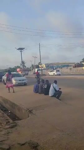 The video shows an outdoor scene on a street corner. Several people are present. A group of four men sit on a low concrete structure, while another man, dressed in a light-colored traditional outfit, stands nearby, looking away from the camera with his hands behind his back. Cars and a motorcycle pass by on the street. A building with a blue awning is visible in the background, along with power lines overhead. The sky appears overcast.