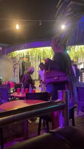 The video shows an outdoor scene at night, likely a bar or restaurant. In the foreground, a person wearing a black dress and carrying a quilted handbag is partially visible, facing away from the camera. Their hair is long and braided. Behind them, a group of people are gathered. To the left, a person with a shaved head wearing a dark top is seated at a table with red cups. Further back, several individuals are standing and interacting. The environment includes tables and chairs, string lights, and tropical plants. Blue and red lighting illuminates the area.