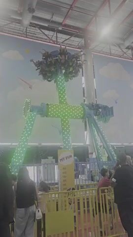 The video shows an amusement park ride with people on it. The ride is tall and has lights that change color. There are people standing in a line waiting to ride. The background is a mural of clouds and airplanes. The lighting is bright.