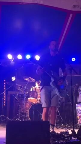 The video captures a concert scene illuminated by blue stage lights. In the foreground, a person holding a light-colored electric guitar stands on a stage, wearing a dark t-shirt, denim shorts, and checkered sneakers. The person is singing into a microphone and appears to be a performer. Behind them, a drum set is visible, along with other stage equipment. The background is largely obscured by darkness and stage lighting, with some indistinct figures and structures suggesting a venue. The camera briefly pans to show the audience or another part of the venue, with blurred figures and purple lighting, before returning focus to the performer. The overall atmosphere is that of a live music performance.